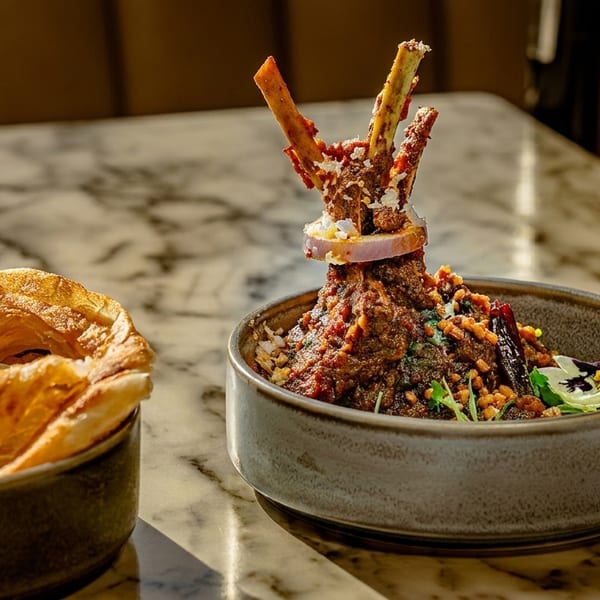 A bowl of lamb shank curry with bone-in shanks, onion, and a side of flaky paratha.